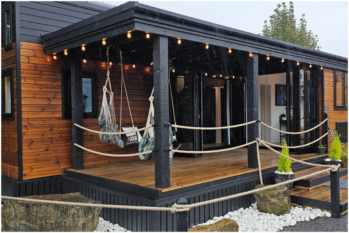 A cozy and stylish mother-in-law suite, possibly a detached tiny home or houseboat, featuring a warm wooden exterior contrasted with black trim, a welcoming covered porch with string lights and hanging chairs, and a small landscaped area with white stones and potted plants.