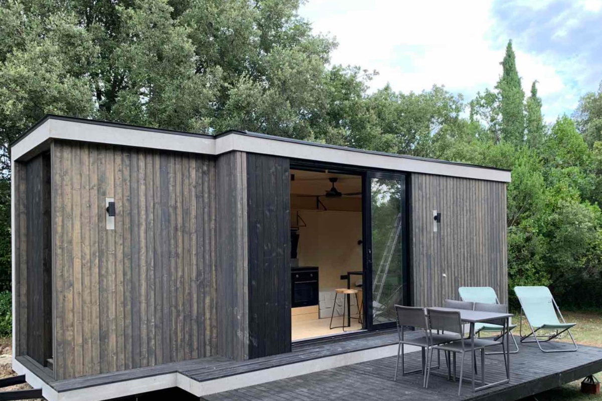 A modern, cube-style detached mother-in-law suite or tiny home with dark wooden siding, a flat roof, large sliding glass doors revealing a minimalist interior, and an outdoor deck area with a table and chairs, set amidst a natural, treed landscape.