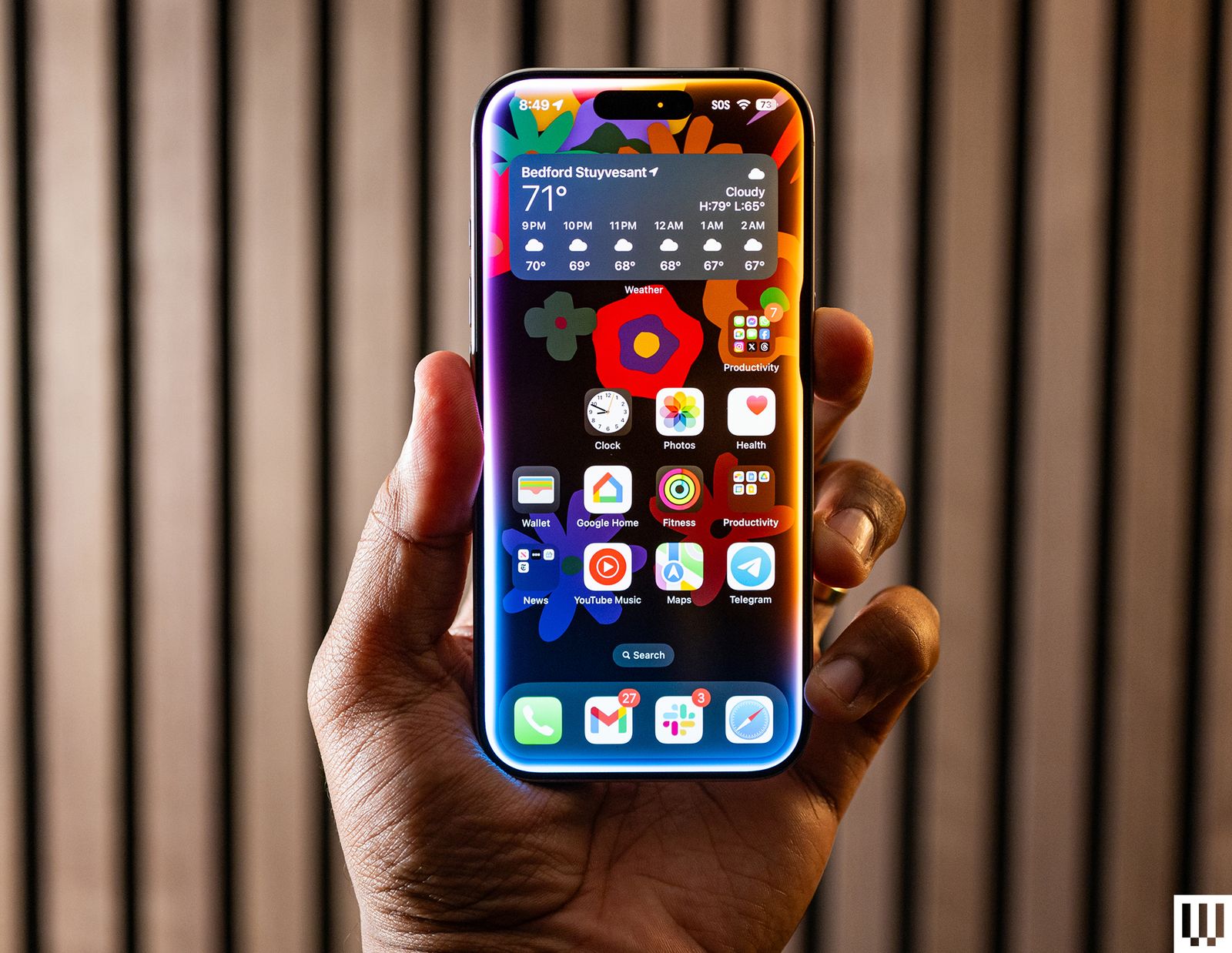 Hand holding an iPhone 16 Pro with the screen showing app icons weather and decorative floral background